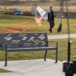 A gray metal park bench sits on a sidewalk with grass and young trees nearby; a person in the background carries a large model airplane.