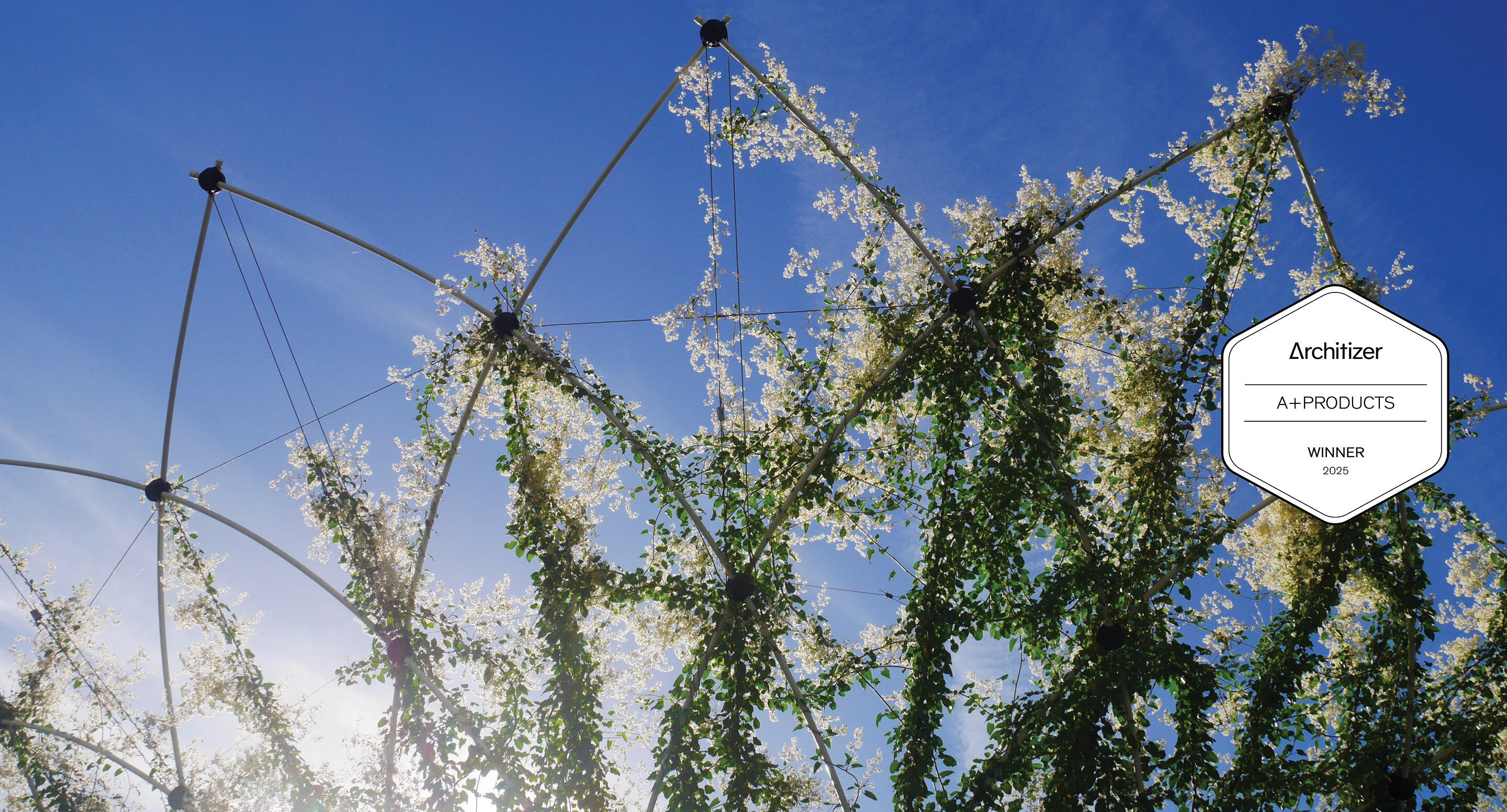 Structure métallique géométrique recouverte de vignes vertes feuillues et de fleurs blanches, sur fond de ciel bleu clair. Un badge indique 