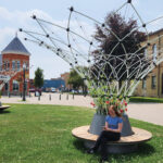 A young woman sits on a circular Ogden bench beneath a large Maglin Corolle - a trellised shade structure - in a grassy plaza with historic brick buildings in the background. A white badge on the image reads 