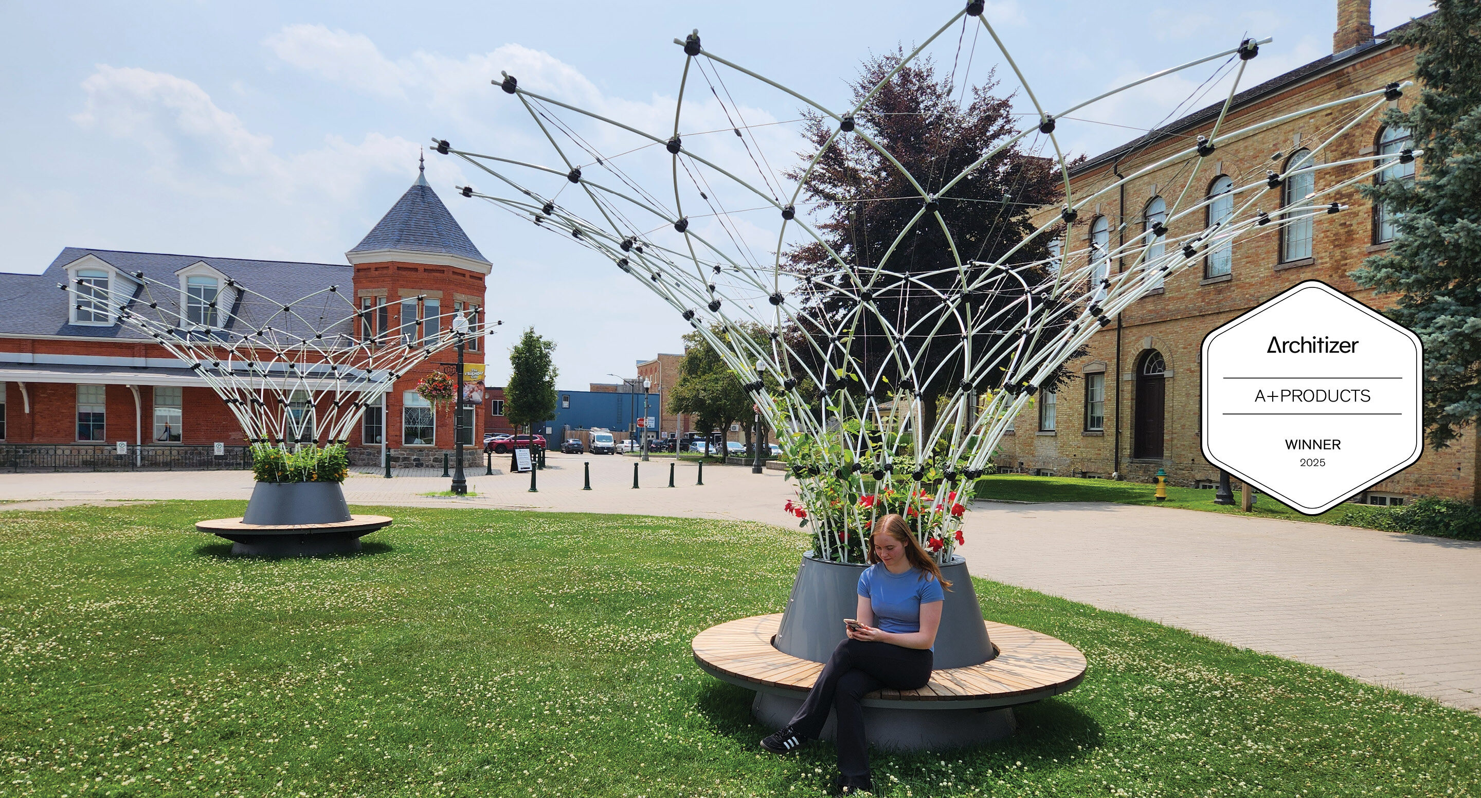 A young woman sits on a circular Ogden bench beneath a large Maglin Corolle - a trellised shade structure - in a grassy plaza with historic brick buildings in the background. A white badge on the image reads 