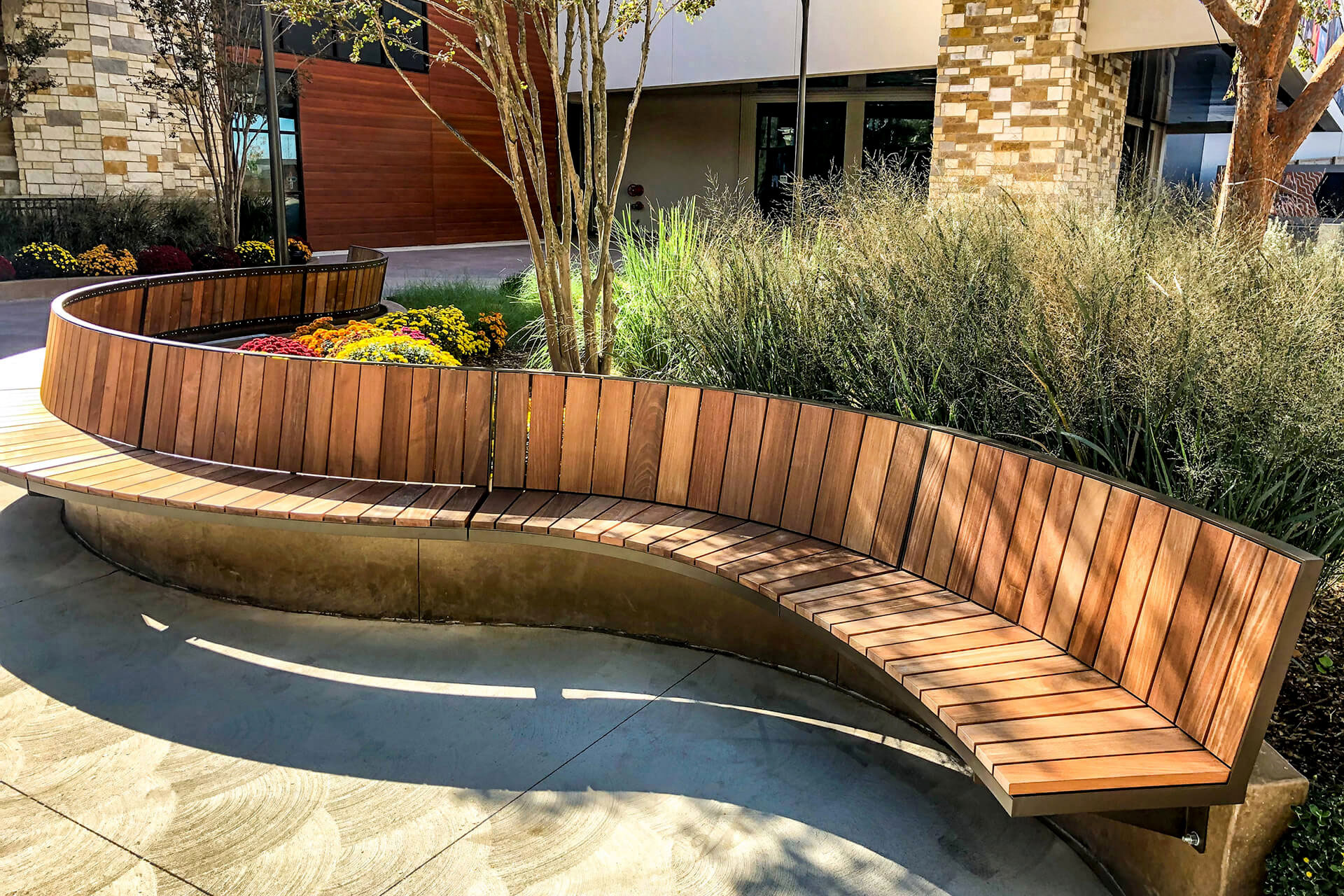 Curved Ogden wooden bench by Maglin, a leader in site furniture, with a metal frame situated outdoors beside landscaping with ornamental grasses and flowers.
