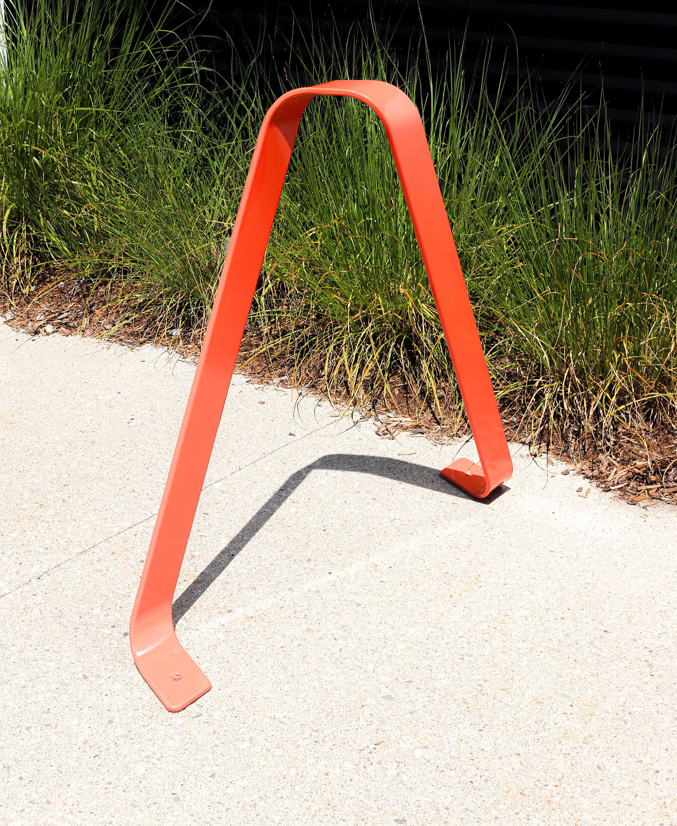 A red metal bike rack shaped like an inverted "V" is installed on a concrete sidewalk next to green grass.