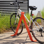 A white bicycle is parked and locked to a bright orange bike rack on a sidewalk near some green grass and striped metal siding.