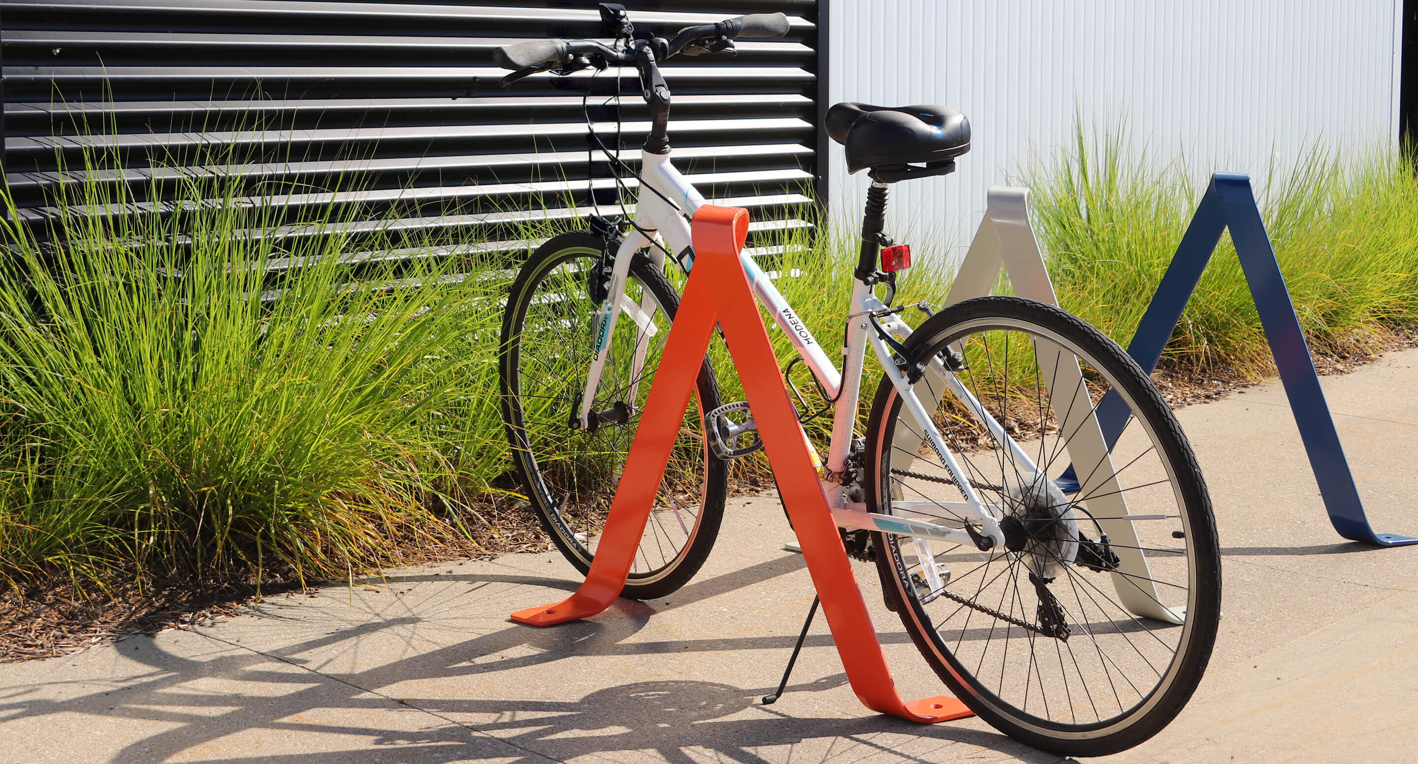 A white bicycle is parked and locked to a bright orange bike rack on a sidewalk near some green grass and striped metal siding.