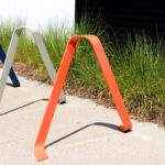 Three metal bike racks, colored blue, white, and orange, are installed on a sidewalk next to tall grass and a building wall.