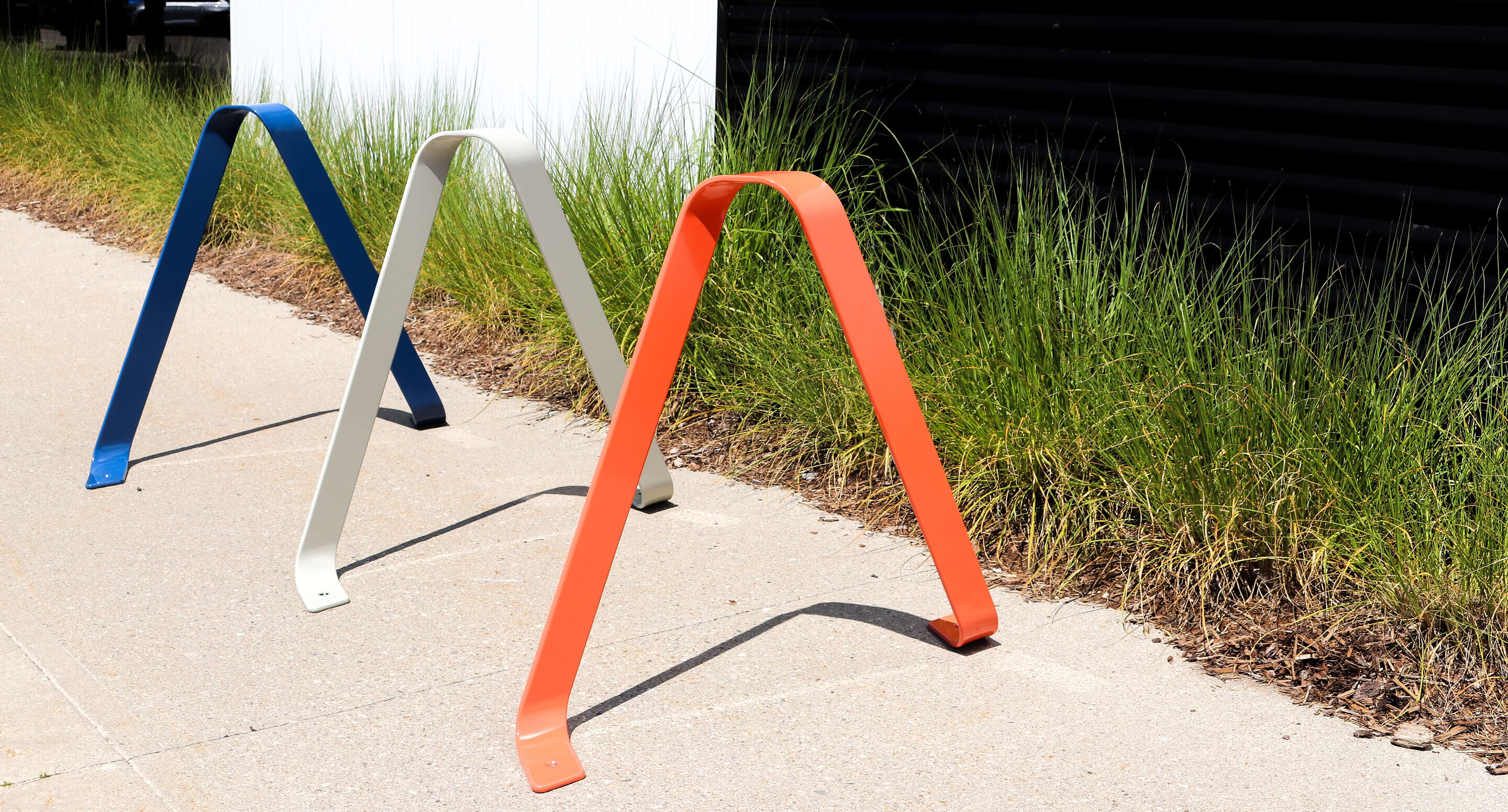 maglin_mbr-3100-bike-racks_carousel2 Three metal bike racks, colored blue, white, and orange, are installed on a sidewalk next to tall grass and a building wall.