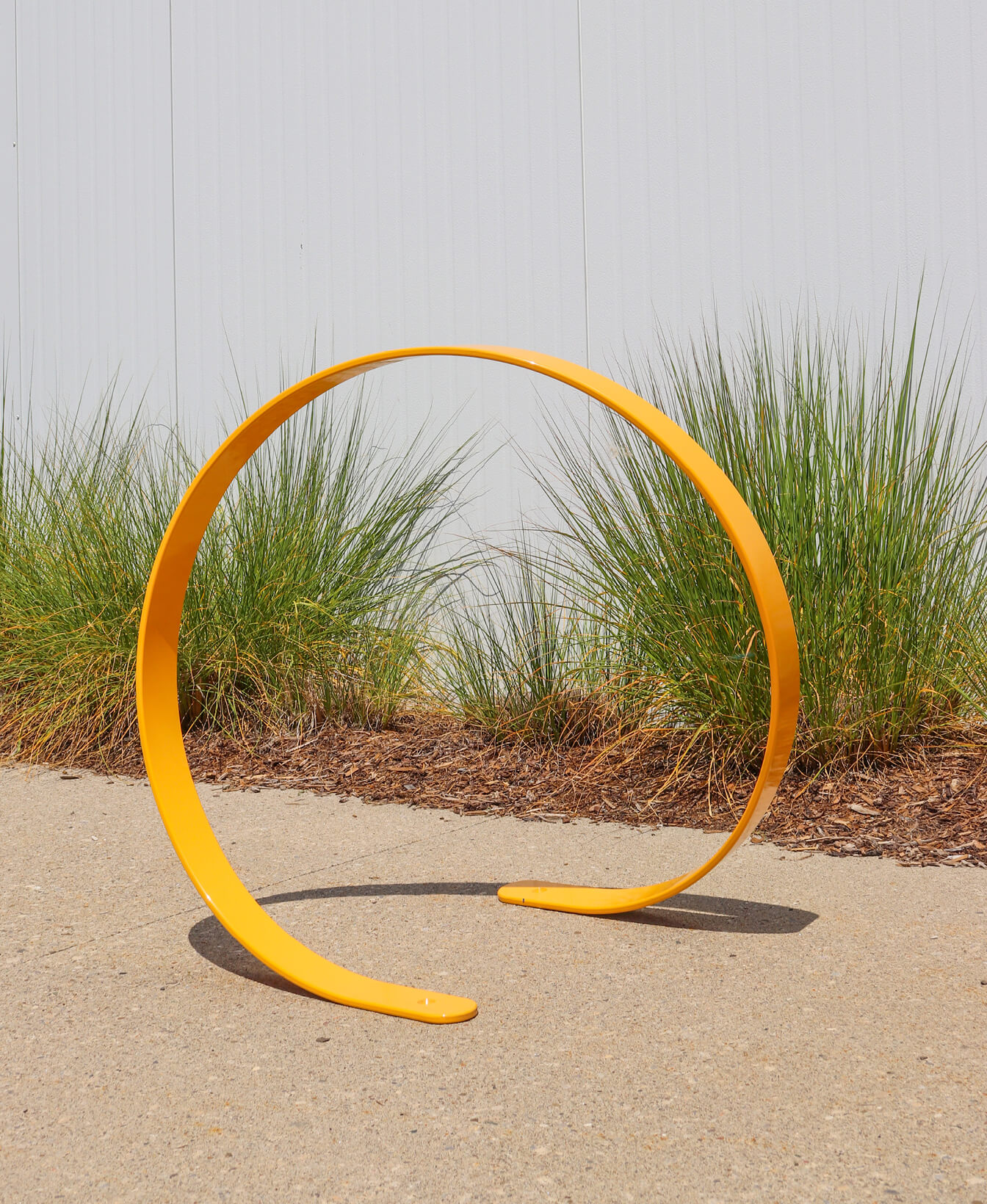 A yellow circular metal bike rack is installed on a concrete sidewalk with ornamental grass and mulch in the background.