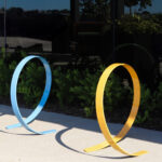 Three colorful metal bike racks, shaped like large loops in yellow, blue, and green, are installed on a sidewalk beside a garden with green shrubs and a reflective glass building.