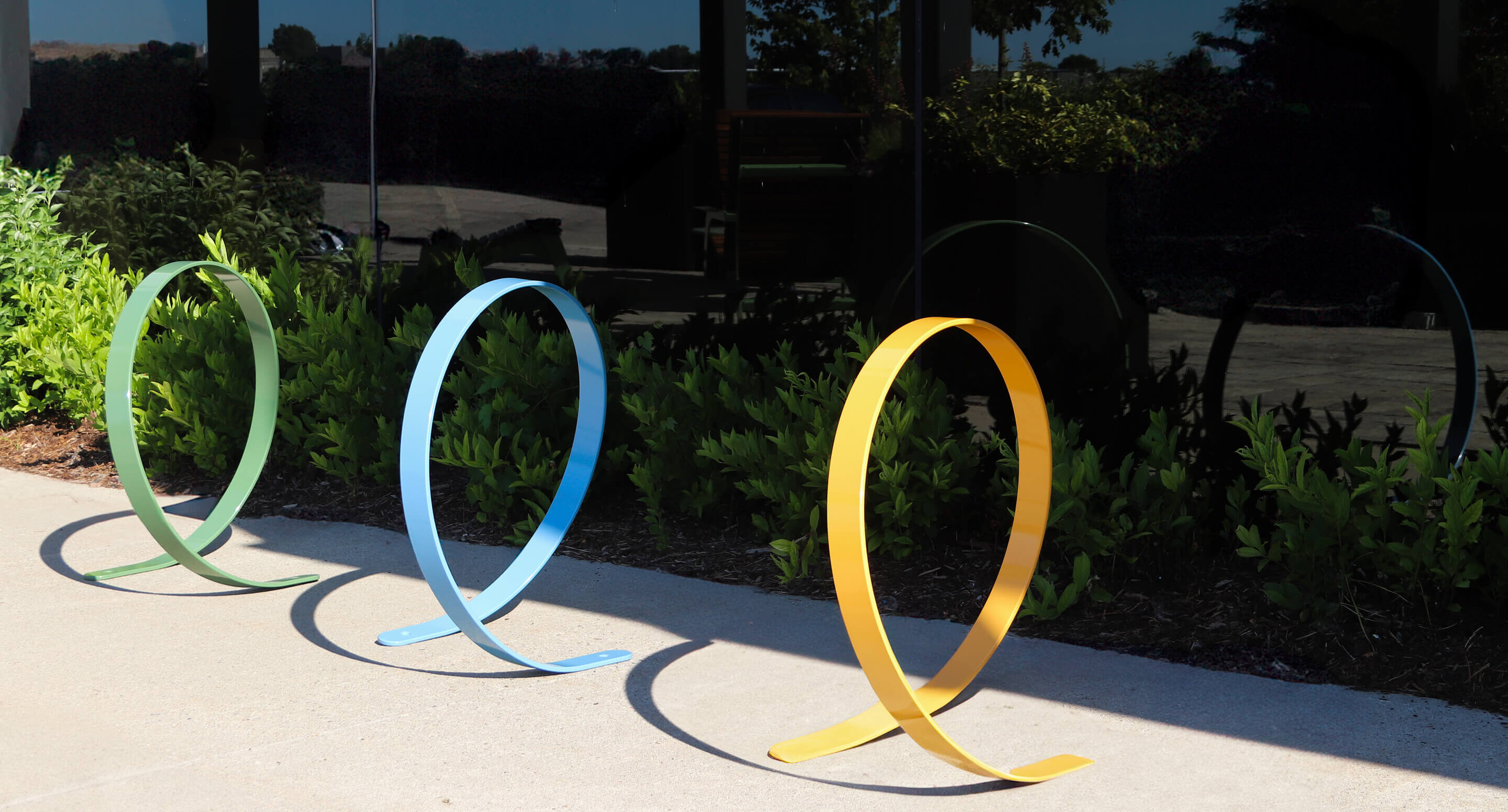 maglin_orbit-bike-racks_carousel Three colorful metal bike racks, shaped like large loops in yellow, blue, and green, are installed on a sidewalk beside a garden with green shrubs and a reflective glass building.