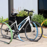 A white bicycle is locked to a curved, colorful bike rack on a paved area near a building with plants and benches nearby.