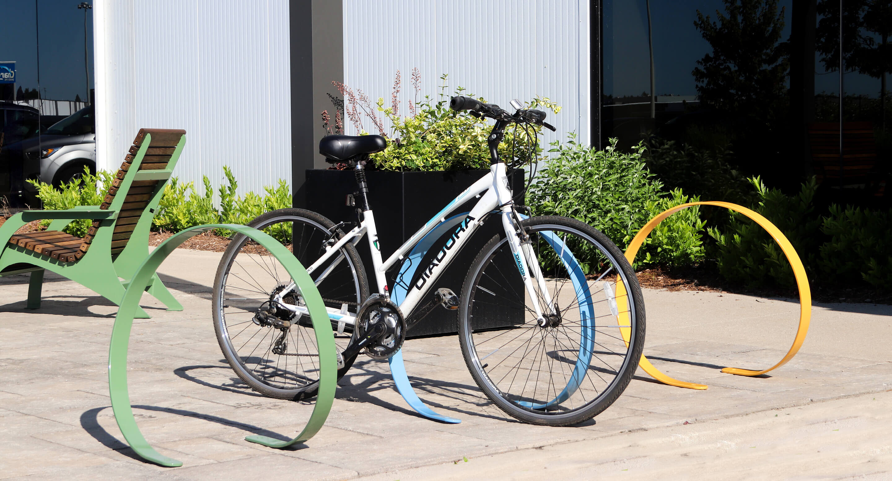 maglin_orbit-bike-racks_carousel2 A white bicycle is locked to a curved, colorful bike rack on a paved area near a building with plants and benches nearby.