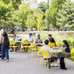 Groups enjoy lunch using movable seating at Battery Tables in Butterfield Park, illustrating the lively campus design and community focus of the space.; trees and green grass surround the area on a sunny day.
