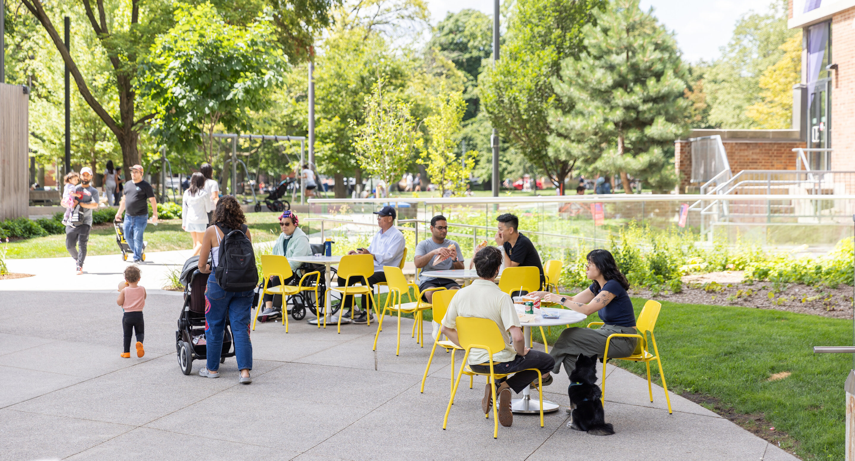 Groups enjoy lunch using movable seating at Battery Tables in Butterfield Park, illustrating the lively campus design and community focus of the space.; trees and green grass surround the area on a sunny day.