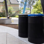 Black 600 Series waste and recycling receptacles in Butterfield Park, complementing the bright movable seating in this vibrant campus design; with benches and trees in the background.