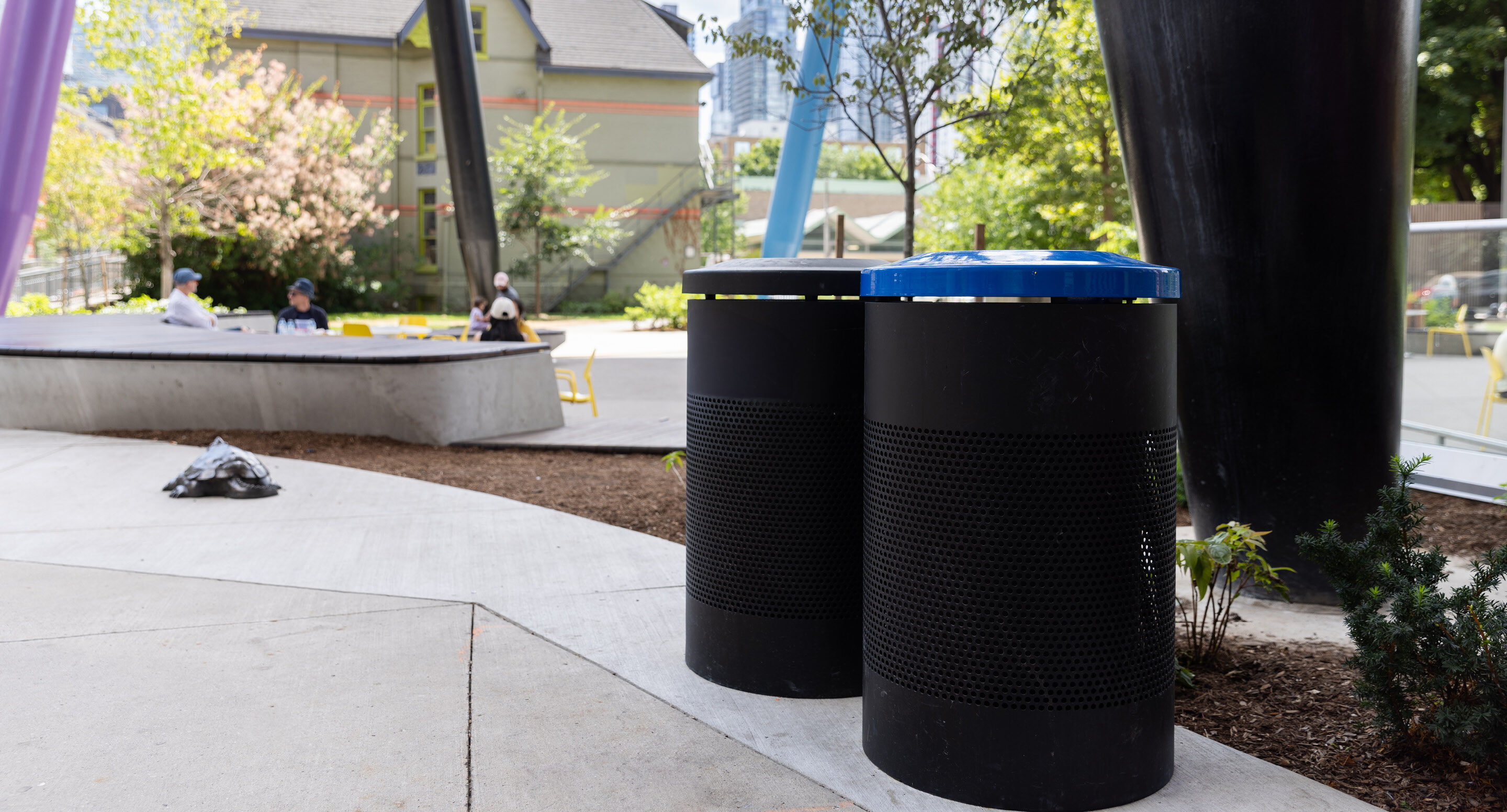 Black 600 Series waste and recycling receptacles in Butterfield Park, complementing the bright movable seating in this vibrant campus design; with benches and trees in the background.