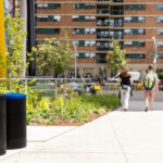 600 Series waste and recycling receptacles positioned near Battery Tables and Aries Café Chairs, supporting sustainability in the campus design of Butterfield Park. Two people walk on a sidewalk next to a colorful mural and modern apartment buildings, with trash bins and greenery along the path.