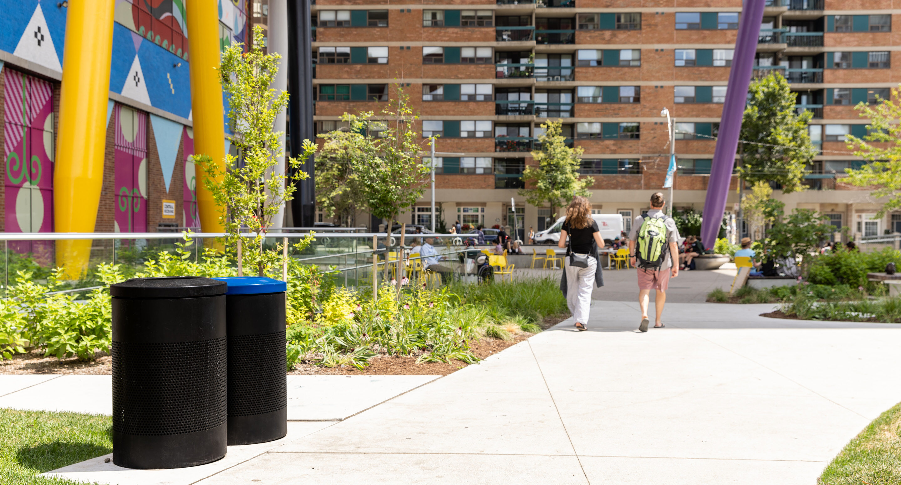 600 Series waste and recycling receptacles positioned near Battery Tables and Aries Café Chairs, supporting sustainability in the campus design of Butterfield Park. Two people walk on a sidewalk next to a colorful mural and modern apartment buildings, with trash bins and greenery along the path.