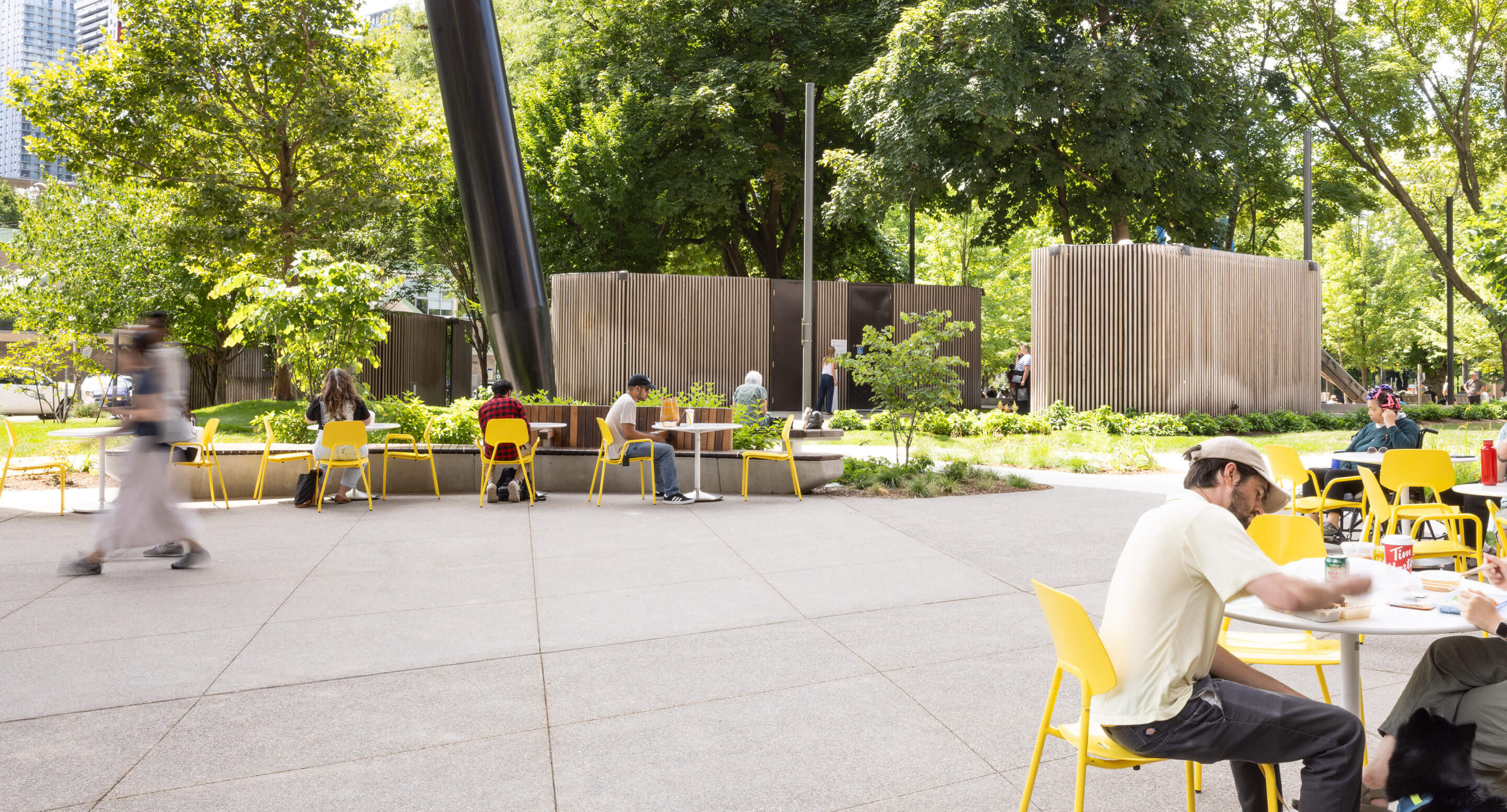 Students and visitors eat lunch, study, and relax at Battery Tables with Aries Café Chairs, showing the social benefits of movable seating in a thoughtful campus design; surrounded by trees and modern structures on a sunny day.