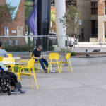 People sit on yellow ARIES Cafe Chairs in an outdoor Butterfield Park at OCAD University, including a person in a wheelchair. Colorful murals and trees are visible in the background.