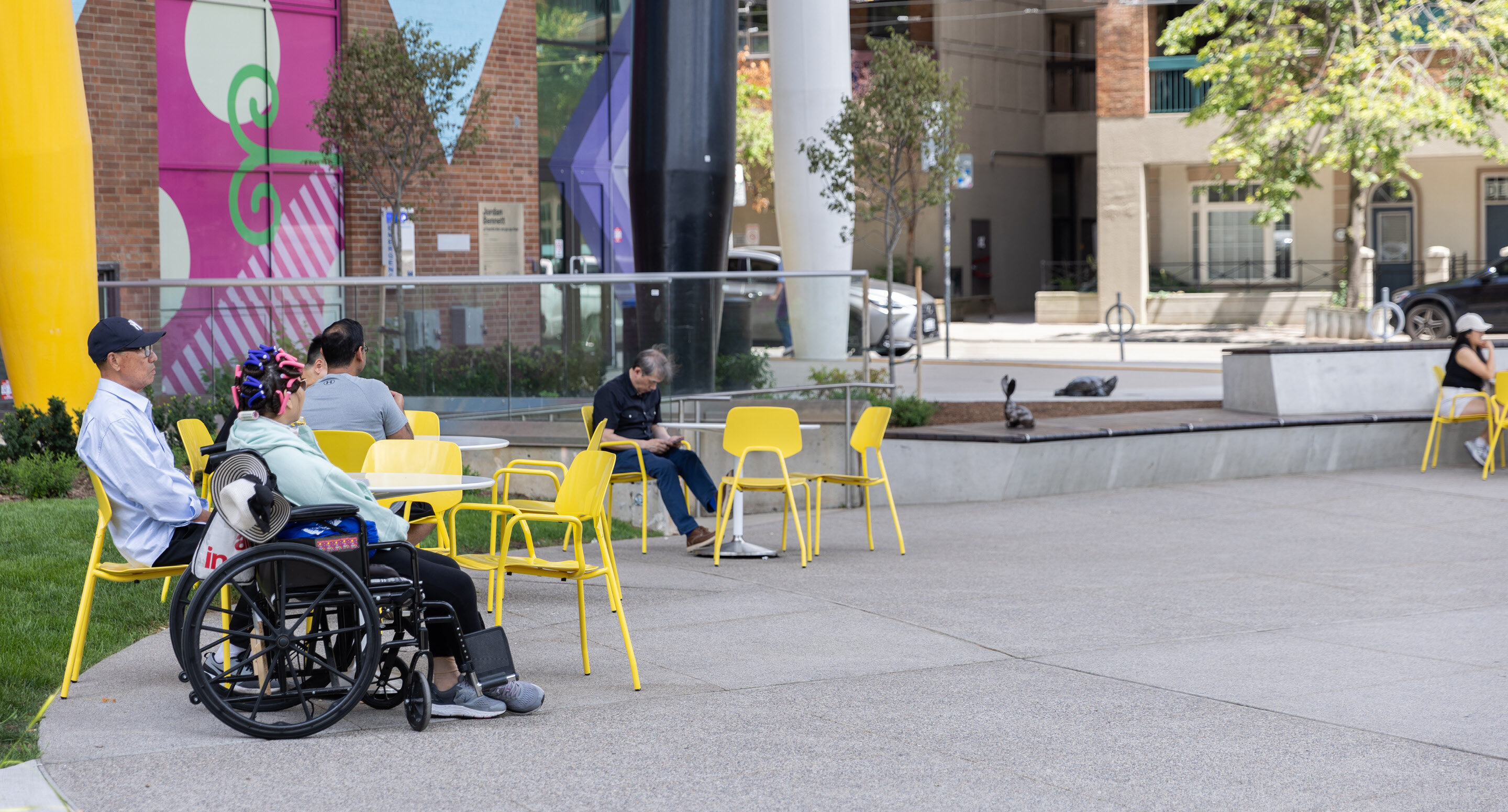 People sit on yellow ARIES Cafe Chairs in an outdoor Butterfield Park at OCAD University, including a person in a wheelchair. Colorful murals and trees are visible in the background.