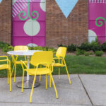 Une table ronde blanche et quatre chaises jaunes sont posées sur un patio en béton devant une peinture murale colorée et un mur de briques, avec de l'herbe et des plantes à proximité.