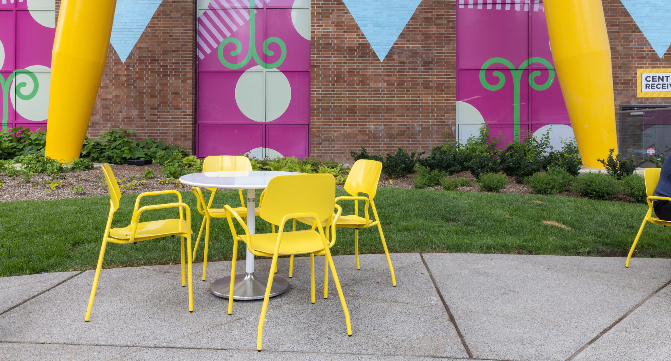 Une table ronde blanche et quatre chaises jaunes sont posées sur un patio en béton devant une peinture murale colorée et un mur de briques, avec de l'herbe et des plantes à proximité.