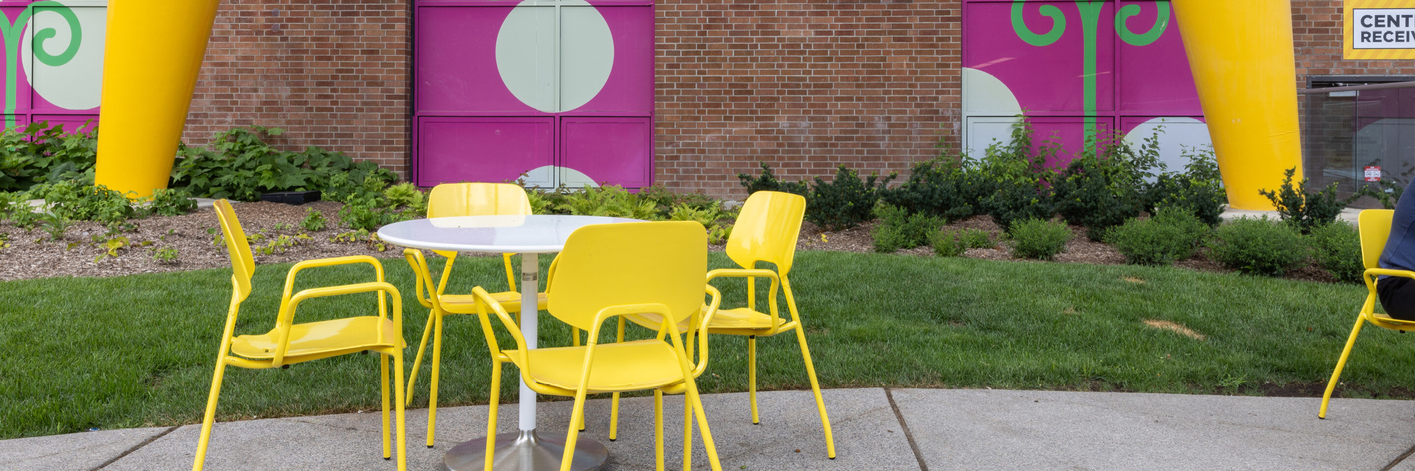 Une table ronde blanche et quatre chaises jaunes sont posées sur un patio en béton devant une peinture murale colorée et un mur de briques, avec de l'herbe et des plantes à proximité.