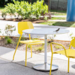 A small bird perches on a Battery Table surrounded by yellow Aries Café Chairs, showing how movable seating enhances the inviting campus design of Butterfield Park.