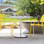 Outdoor seating area with a round white Battery Table and three yellow ARIES Cafe Chairs on a patio, surrounded by greenery and trees, with buildings visible in the background.