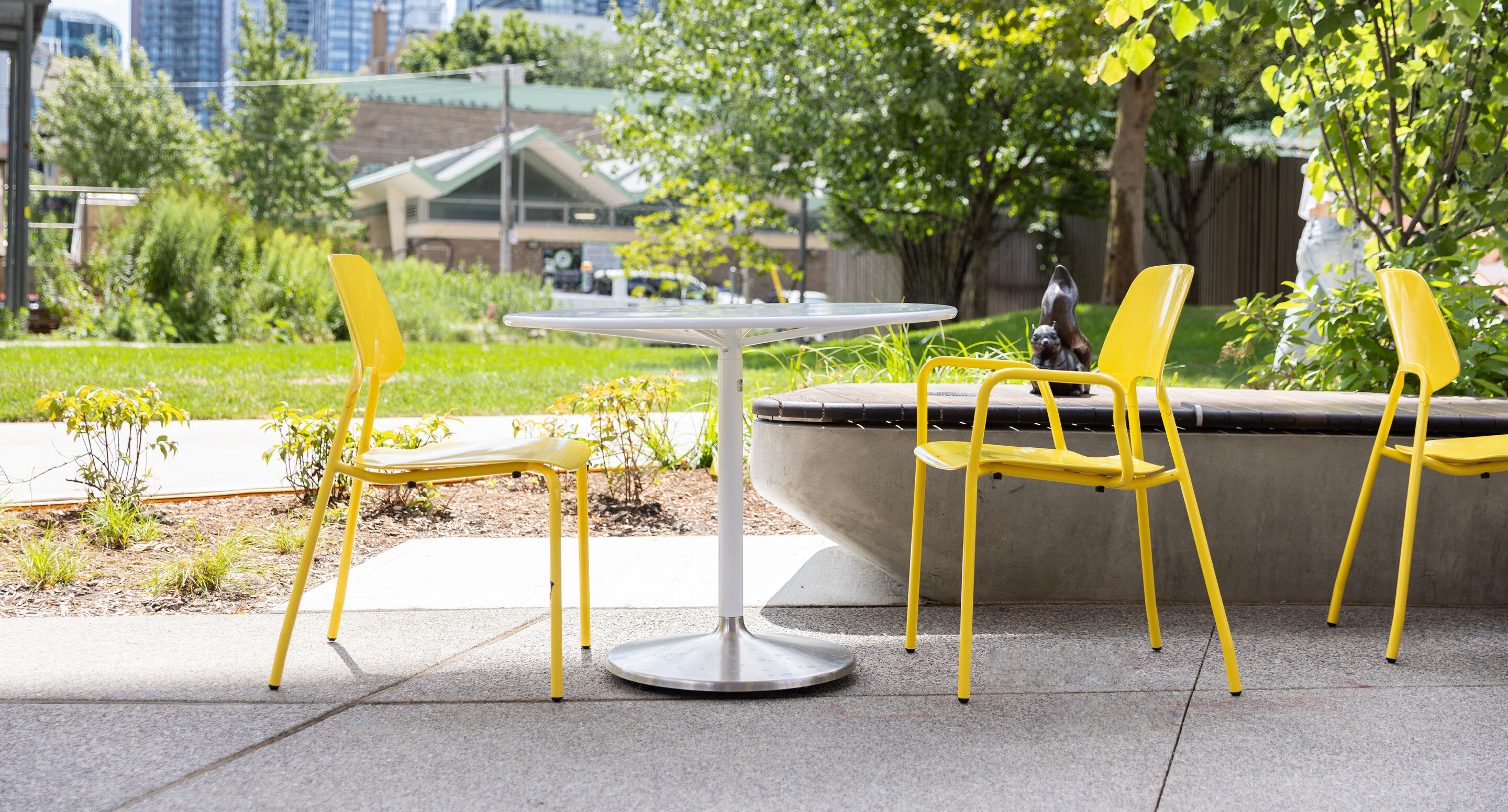Outdoor seating area with a round white Battery Table and three yellow ARIES Cafe Chairs on a patio, surrounded by greenery and trees, with buildings visible in the background.