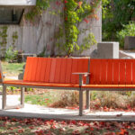 Banc orange incurvé de la collection Ogden avec structure en métal, situé à l'extérieur sur une plate-forme en béton, entouré de feuilles d'automne et de verdure.