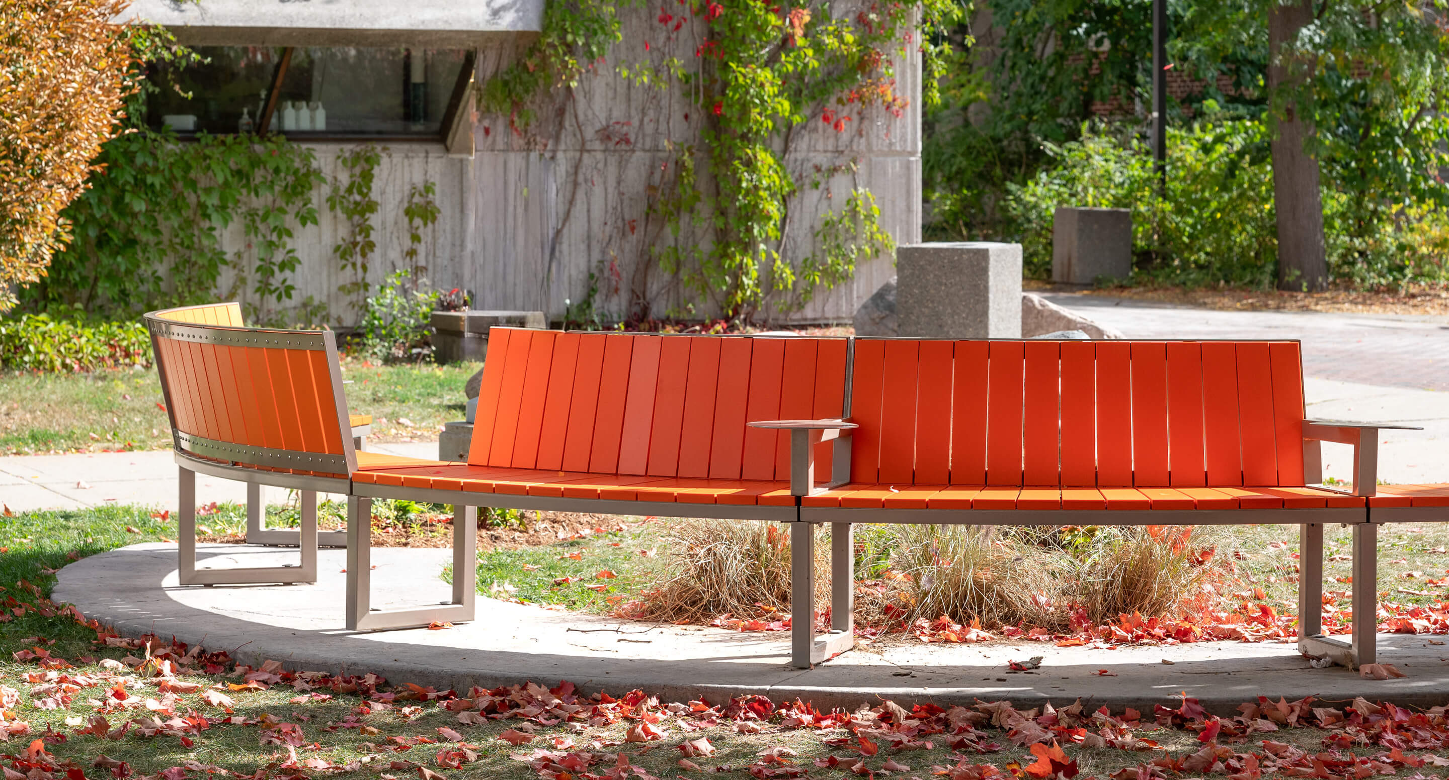 Banc orange incurvé de la collection Ogden avec structure en métal, situé à l'extérieur sur une plate-forme en béton, entouré de feuilles d'automne et de verdure.