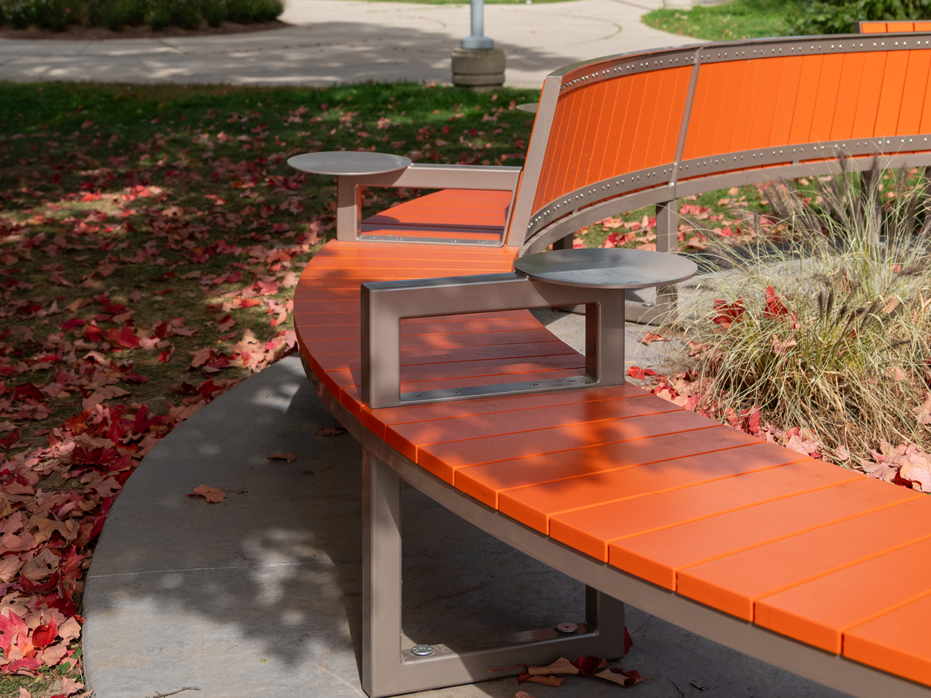 Banc d'extérieur incurvé de la collection Ogden, avec des lattes orange et des tables rondes intégrées, entouré de feuilles rouges tombées et de verdure dans un parc.