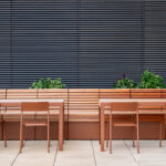 A modern outdoor seating area features brown metal tables and chairs, a long wooden bench, and potted green plants, set against a backdrop of horizontal metal blinds.