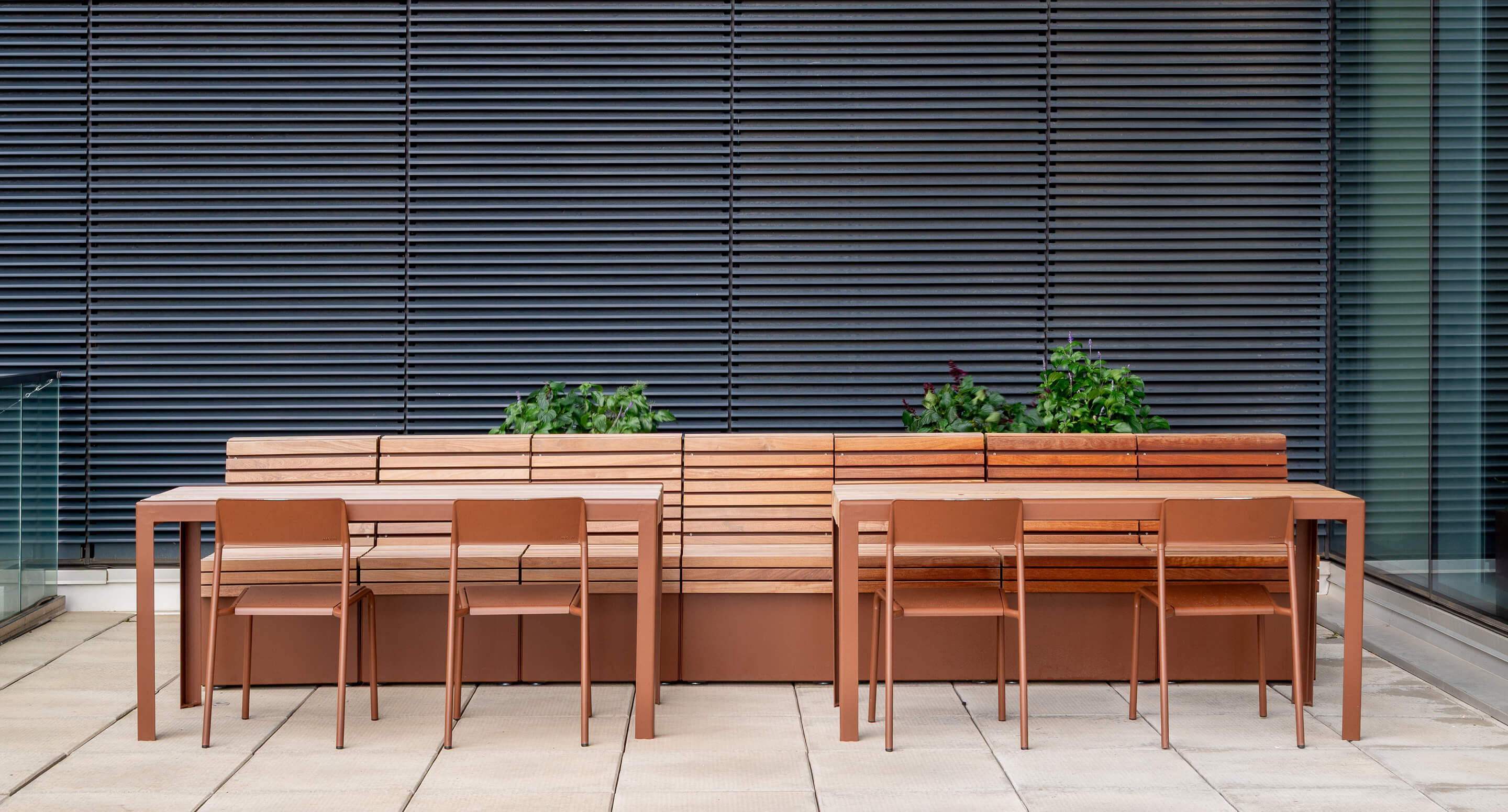 A modern outdoor seating area features brown metal tables and chairs, a long wooden bench, and potted green plants, set against a backdrop of horizontal metal blinds.