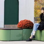 A woman sits on a modern green outdoor bench beside a large potted red chrysanthemum, with autumn foliage and buildings in the background.