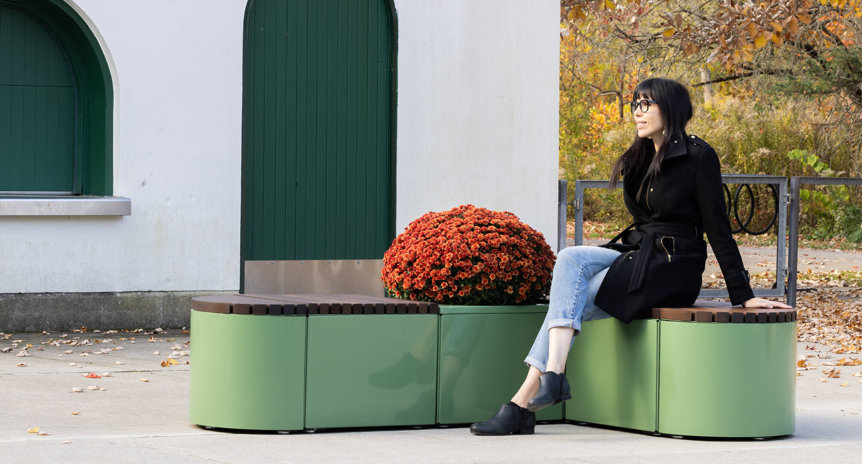 A woman sits on a modern green outdoor bench beside a large potted red chrysanthemum, with autumn foliage and buildings in the background.