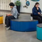 Two people sit on colorful round Pixel benches in a modern indoor space, both looking at their phones; lockers and plants are visible in the background.