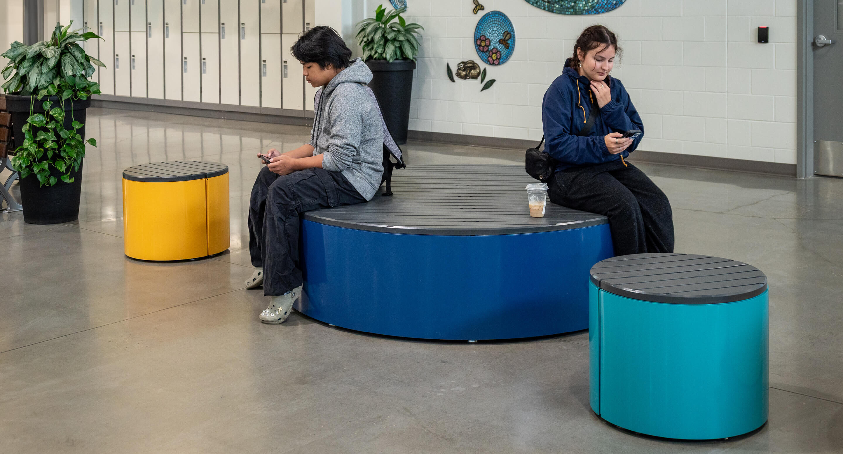Two people sit on colorful round Pixel benches in a modern indoor space, both looking at their phones; lockers and plants are visible in the background.