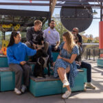 A group of people and two dogs sit and talk on blue benches in an outdoor seating area under a metal canopy on a sunny day.