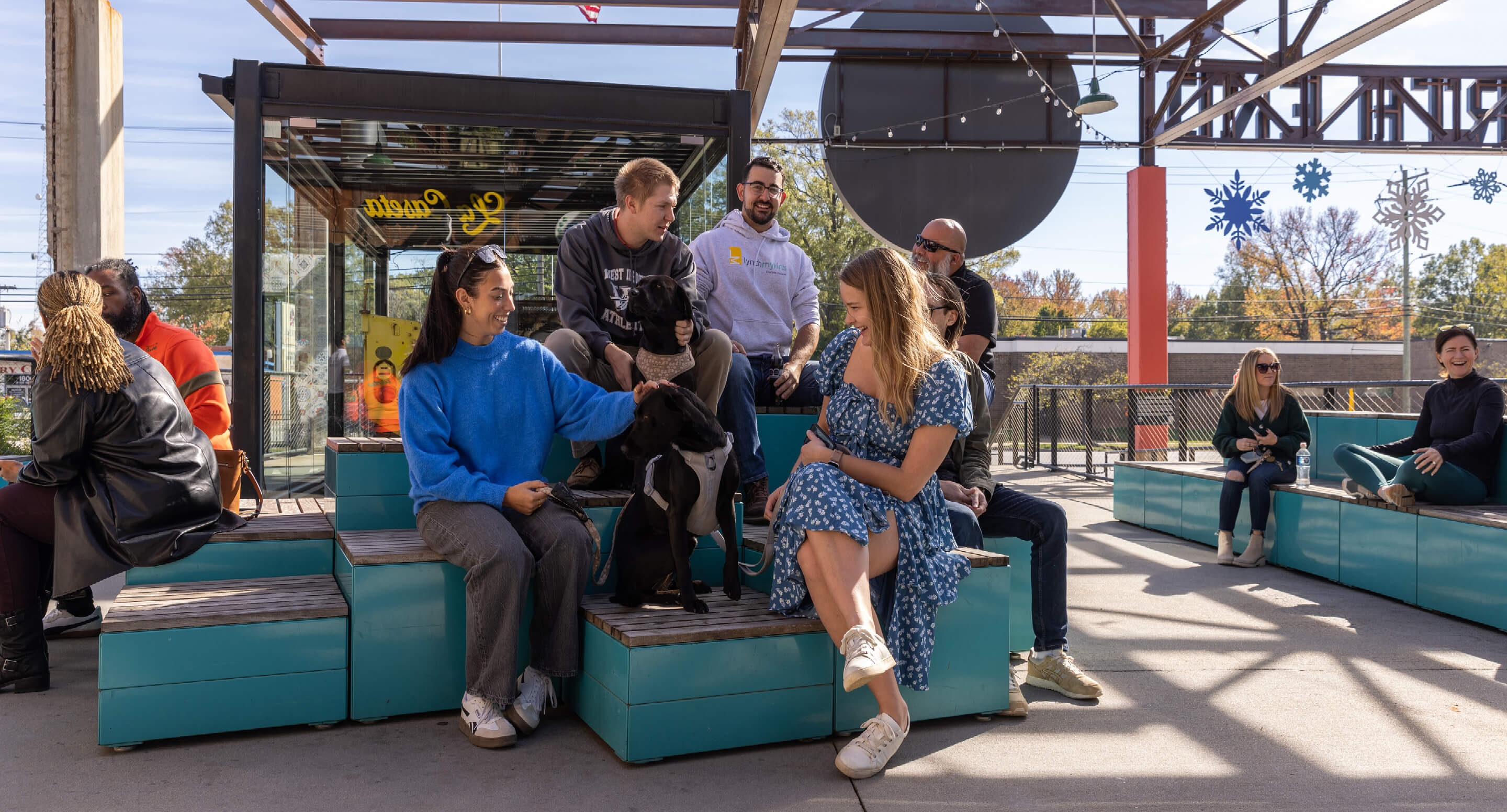 A group of people and two dogs sit and talk on blue benches in an outdoor seating area under a metal canopy on a sunny day.