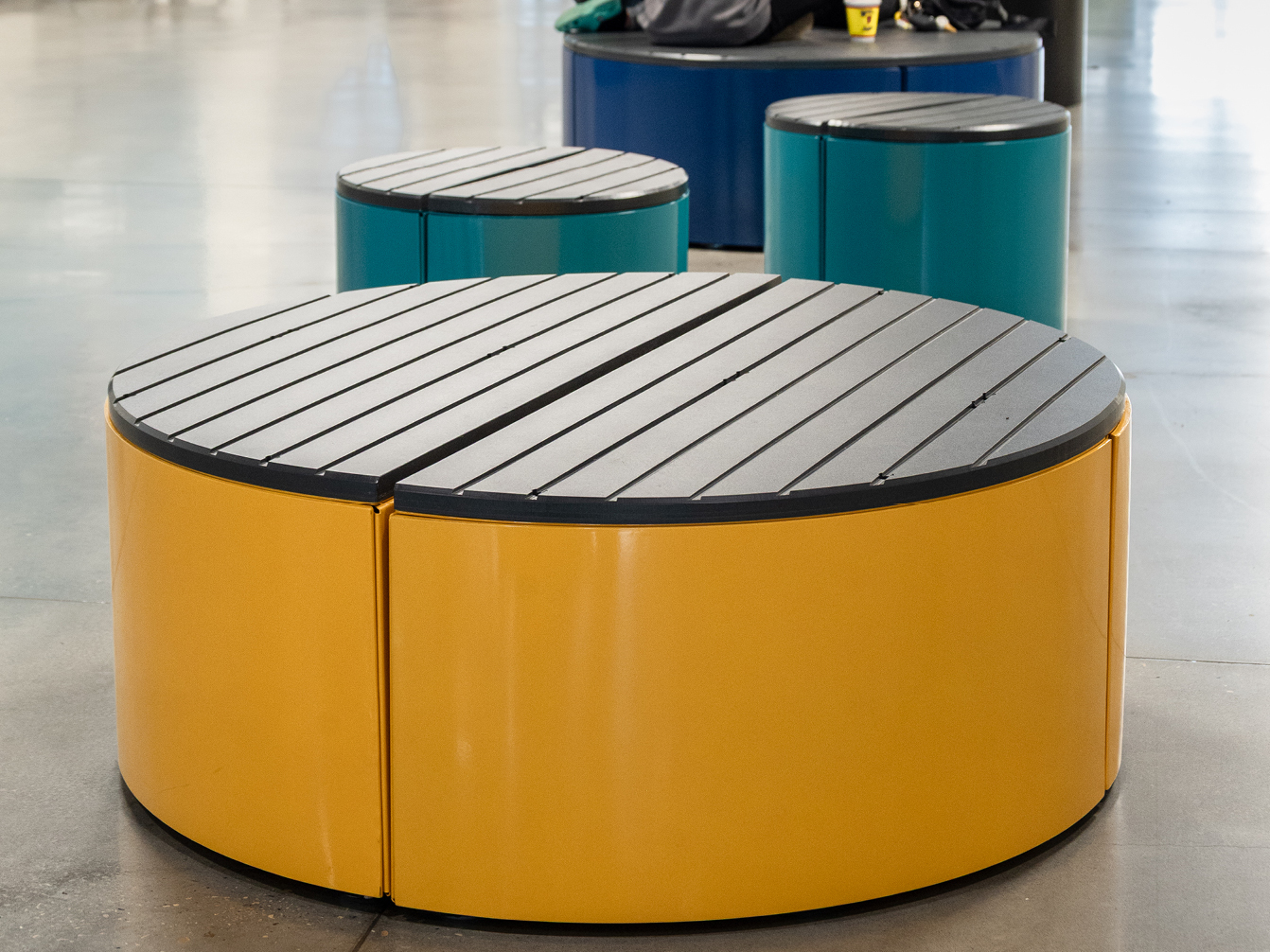 Round, modern benches with slatted black tops and colorful bases in yellow and teal arranged on a polished indoor floor.