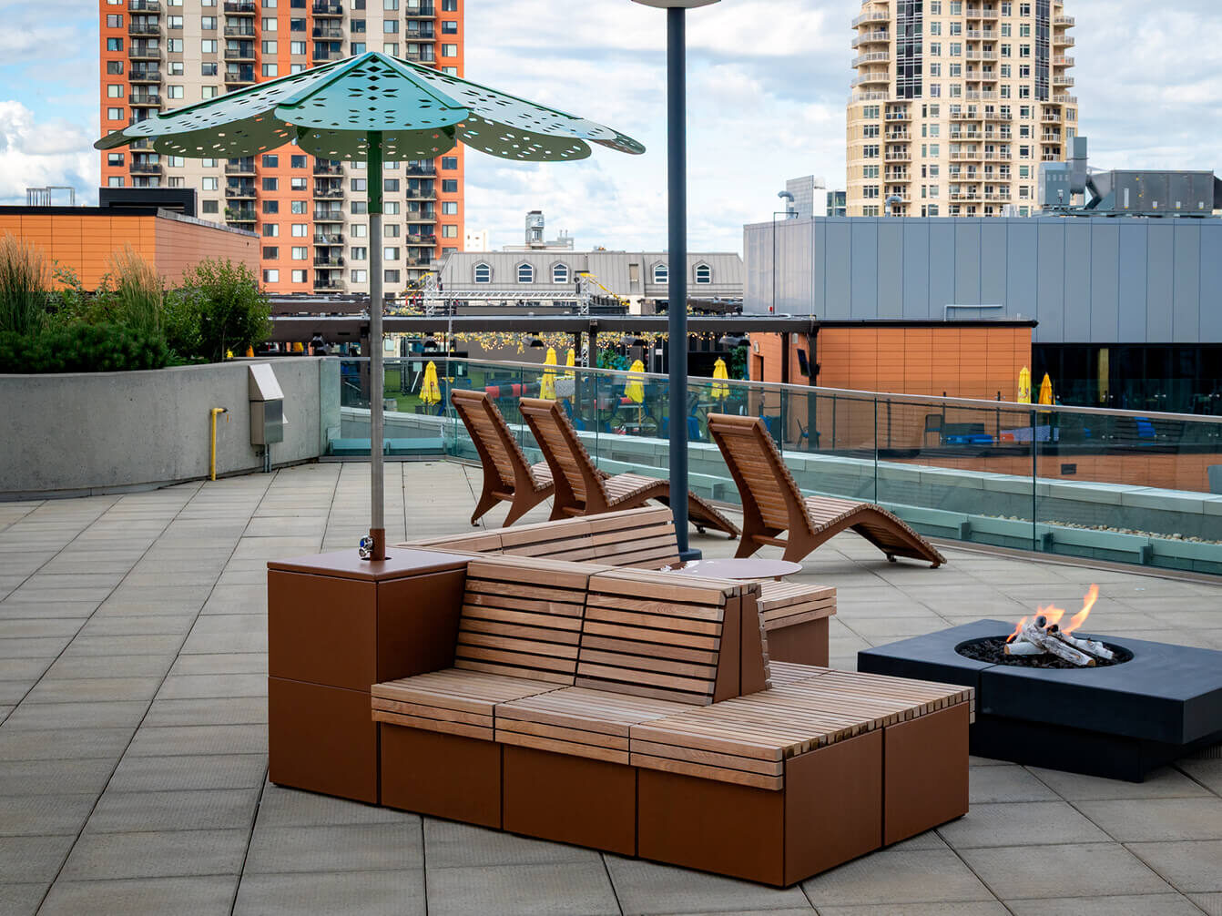 Rooftop patio with wooden lounge chairs, a bench with a sunshade, a fire pit, and city buildings in the background.