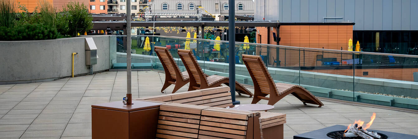 Rooftop patio with wooden lounge chairs, a bench with a sunshade, a fire pit, and city buildings in the background.