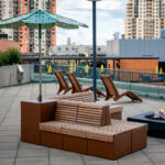 Rooftop patio with wooden lounge chairs, a bench with a sunshade, a fire pit, and city buildings in the background.