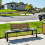 A wooden 1050 bench and a matching trash bin are placed on a sidewalk near a crosswalk, with motorcycles and buildings in the background on a sunny day.