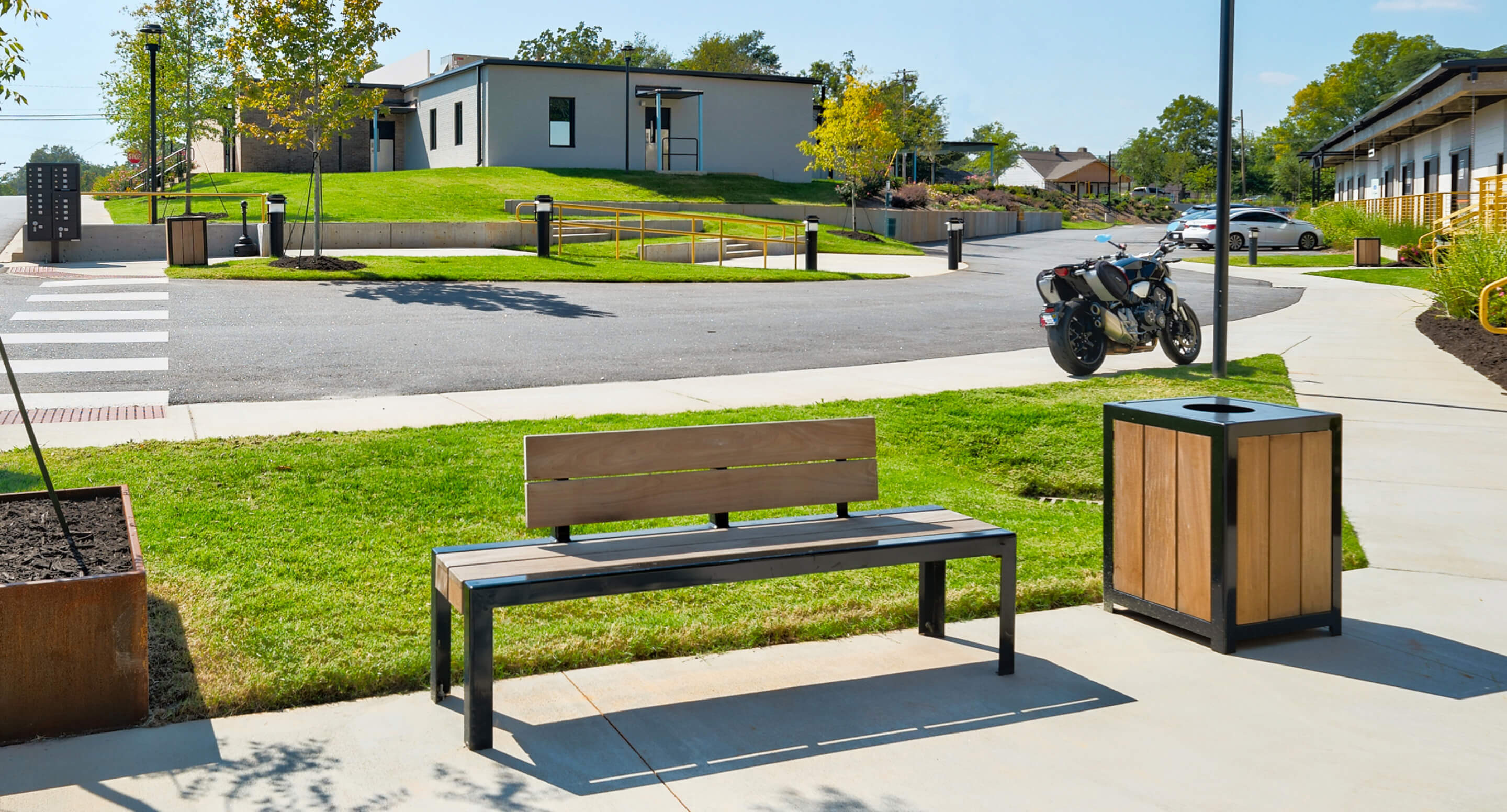A wooden 1050 bench and a matching trash bin are placed on a sidewalk near a crosswalk, with motorcycles and buildings in the background on a sunny day.