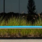 A blue 970 backless hdpe bench sits in front of tall ornamental grass and a dark reflective window.