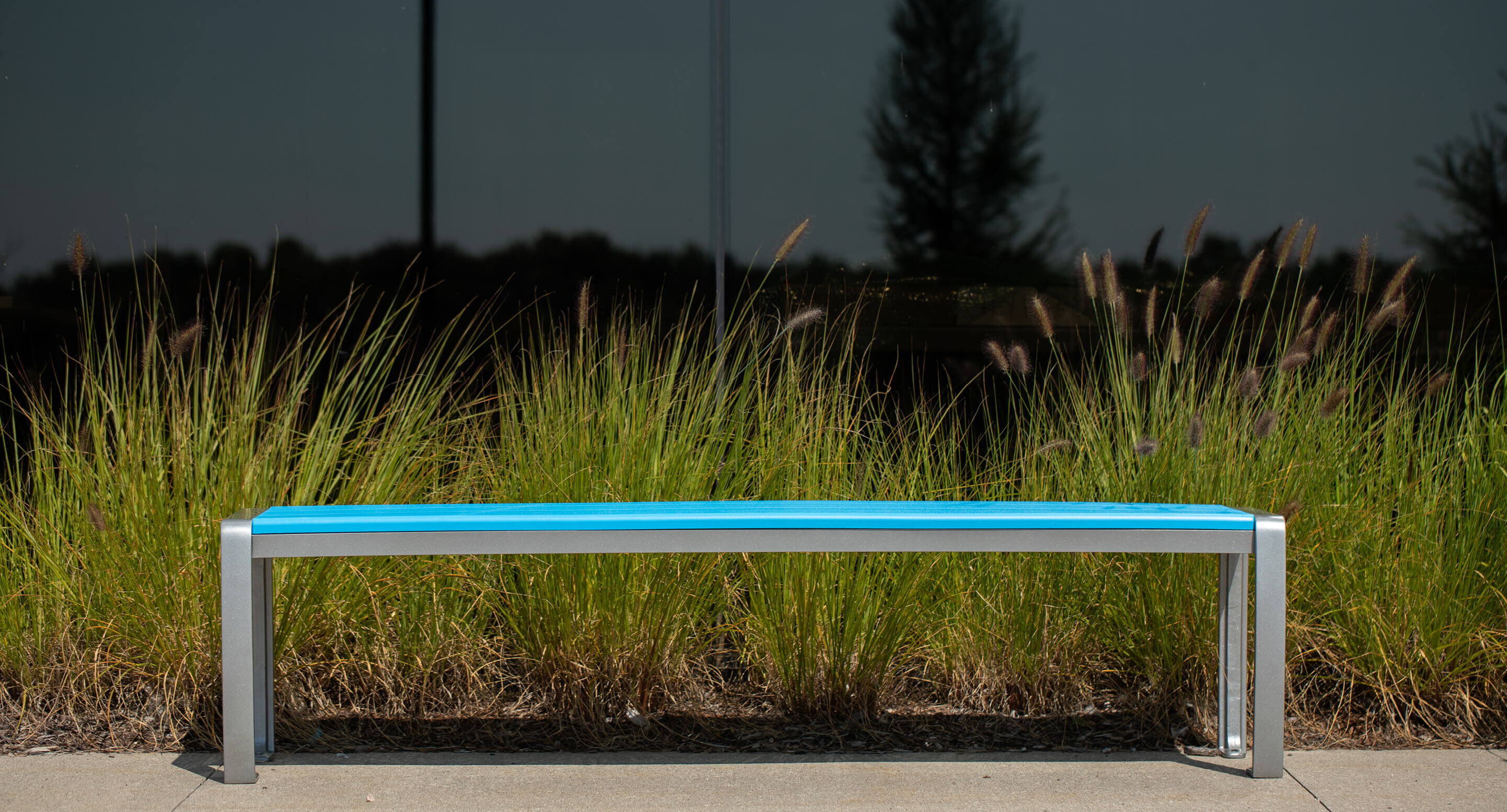 A blue 970 backless hdpe bench sits in front of tall ornamental grass and a dark reflective window.