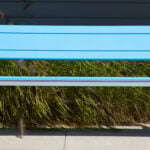 A modern blue 970 bench with metal legs stands on a concrete surface in front of tall grass and a dark building wall.